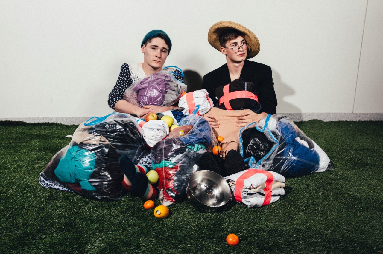 Two male students sitting for a photoshoot surrounded by clothing in plastic bags and fruit